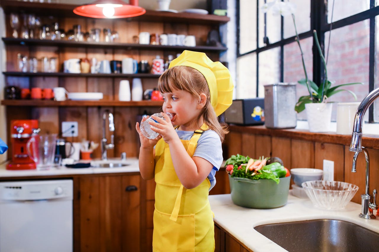 A young girl dressed as a chef drinks water in a cozy kitchen with wooden shelves.